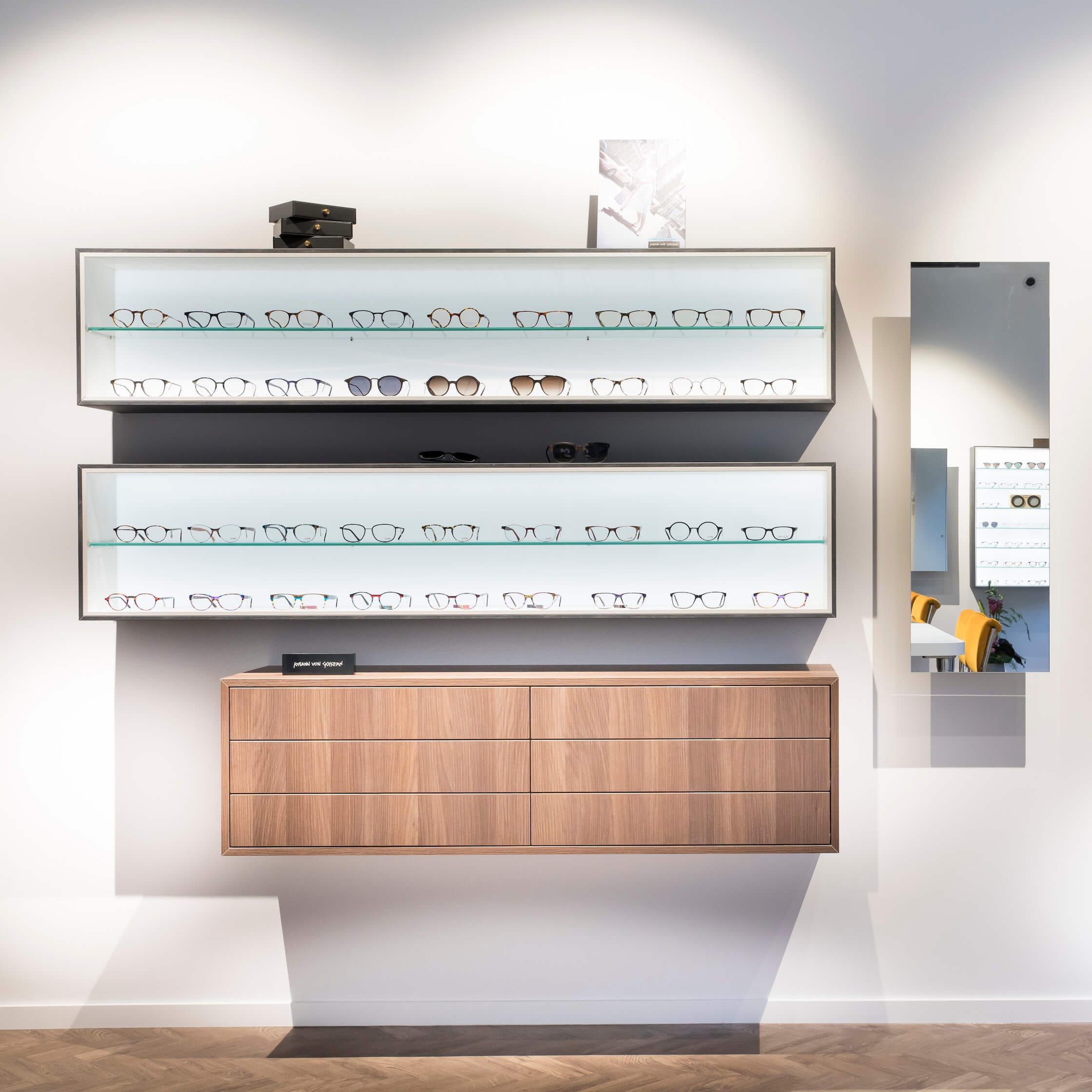 Wall-mounted lit eyewear cases above a walnut cabinet beside a tall mirror