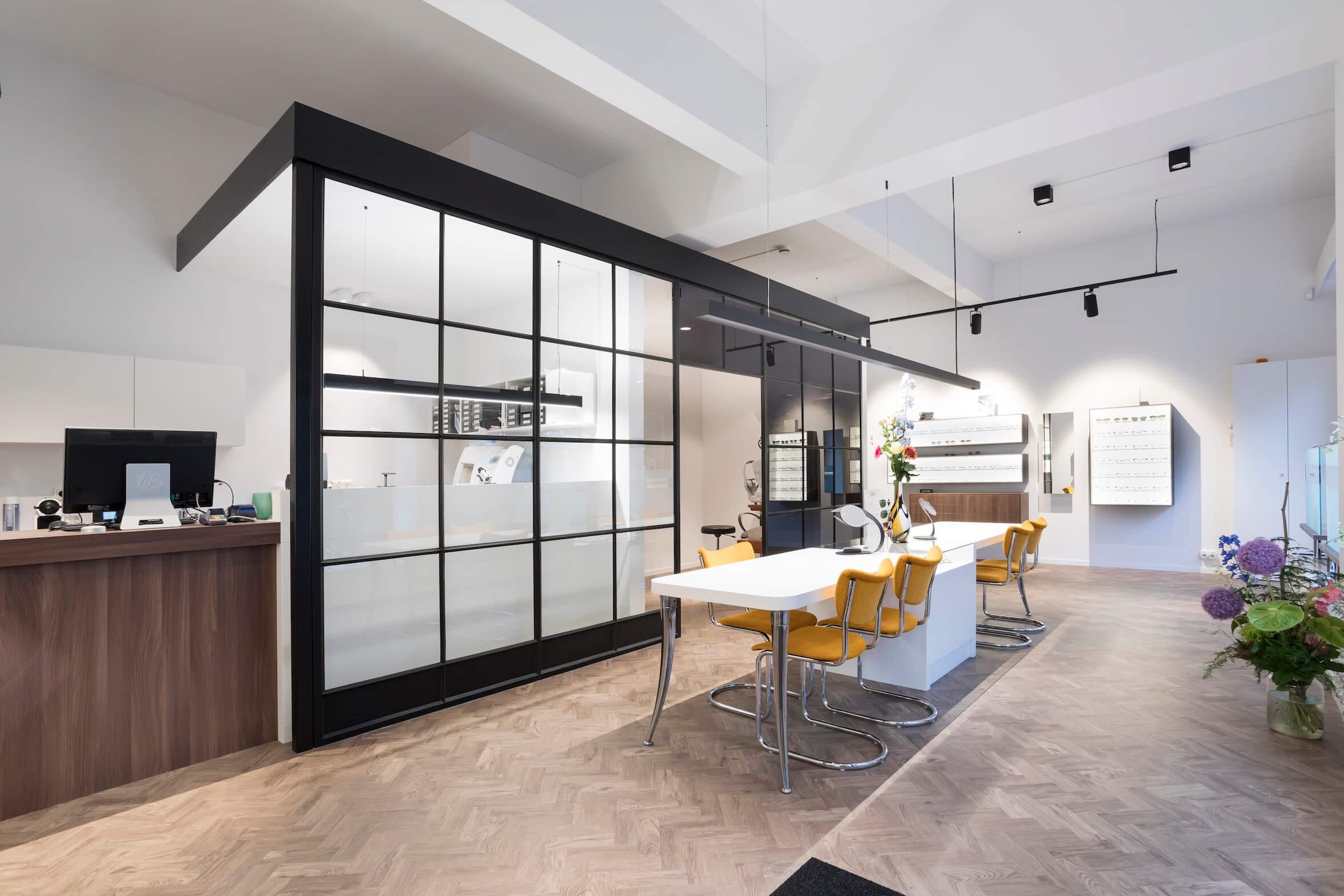 Long white table with yellow chairs in front of a black glazed consultation room