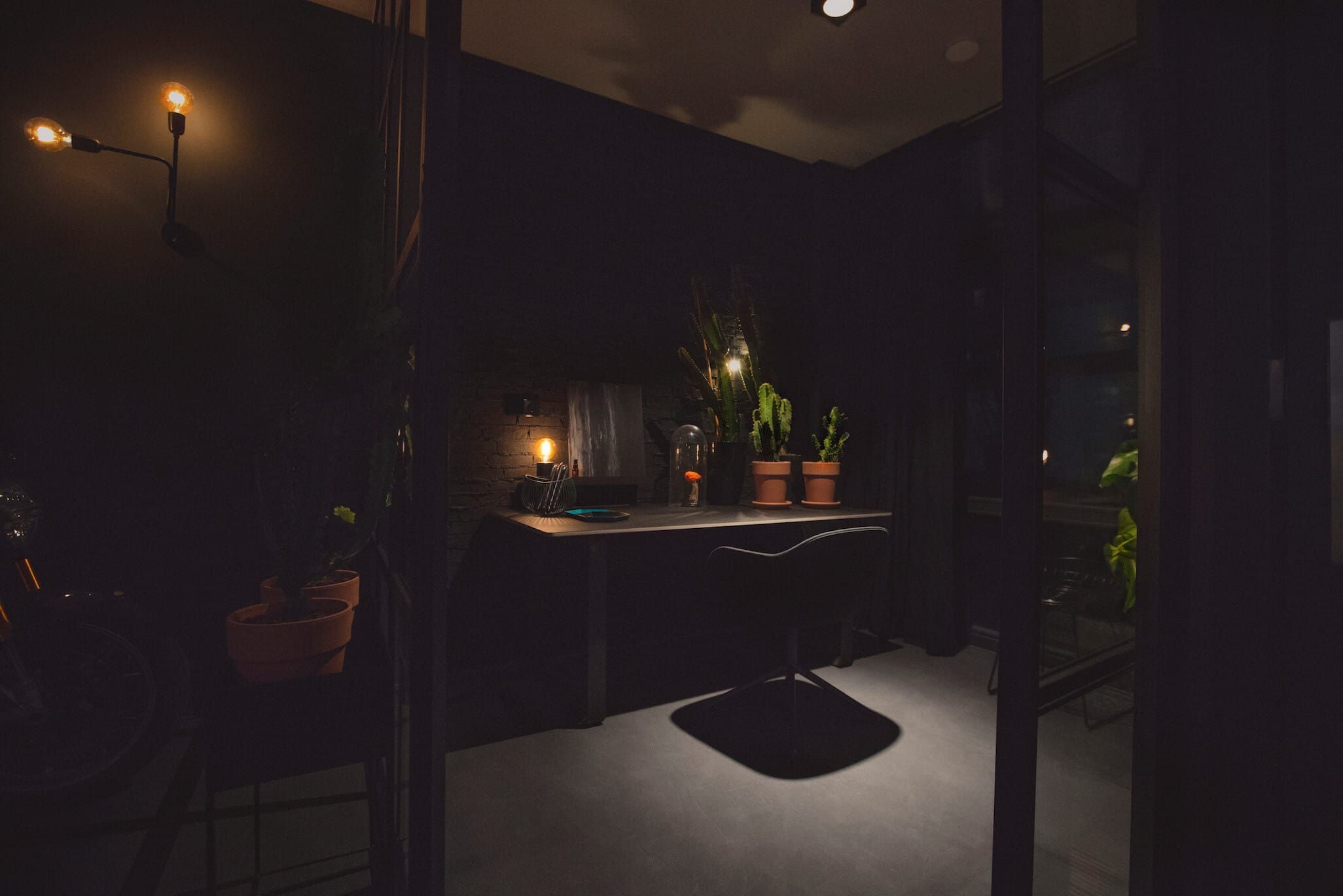 Dark home office nook with desk, plants and warm lamps seen through glass