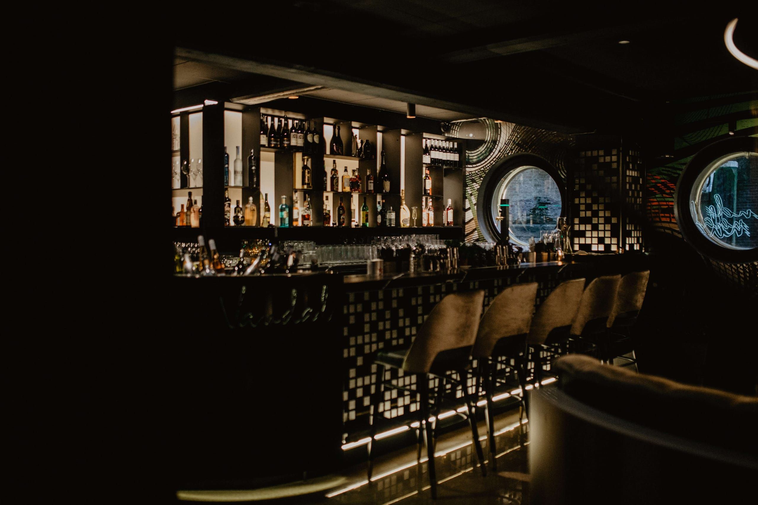 Bar with bottle shelves, mosaic front and velvet stools in low light at Vandal