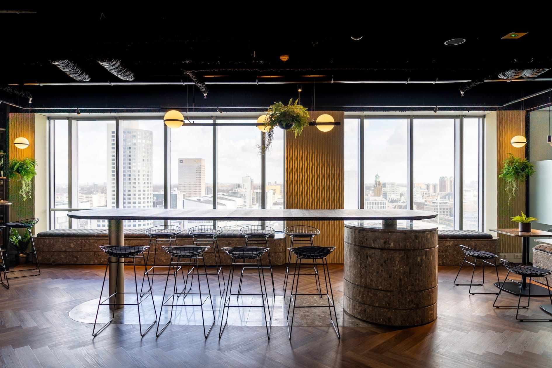 Long high table with wire stools in front of a skyline view and a stone-clad column
