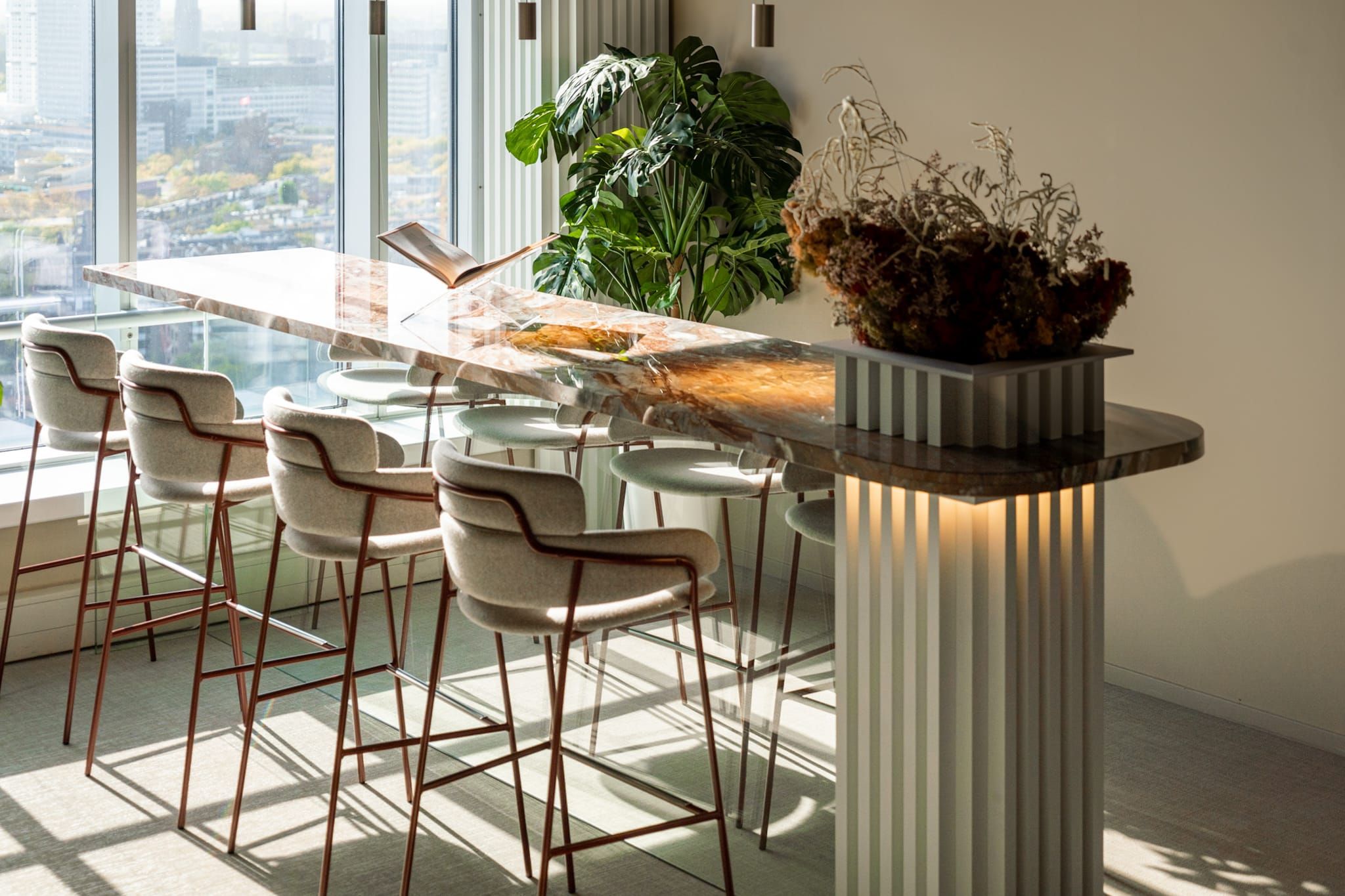 High marble counter with stools in sunlight beside a fluted base and monstera