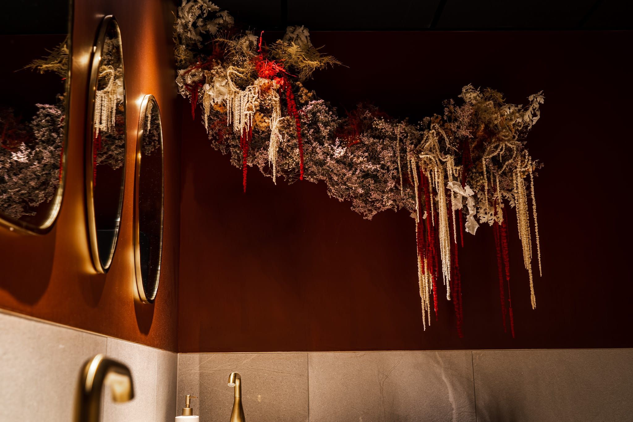 Dried flower installation above round mirrors and brass taps in a washroom