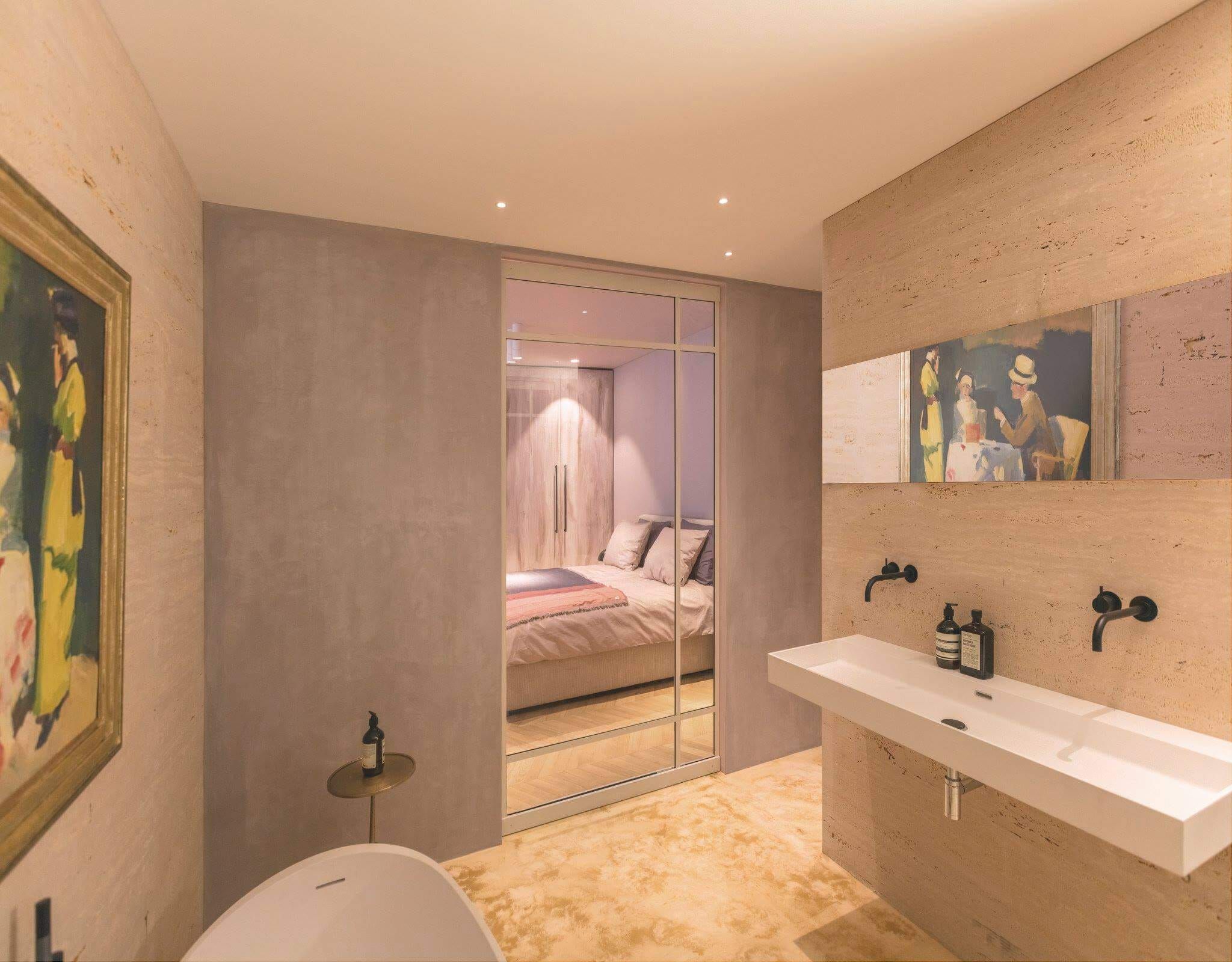 Bathroom with long white basin, black taps and a bedroom visible through glass
