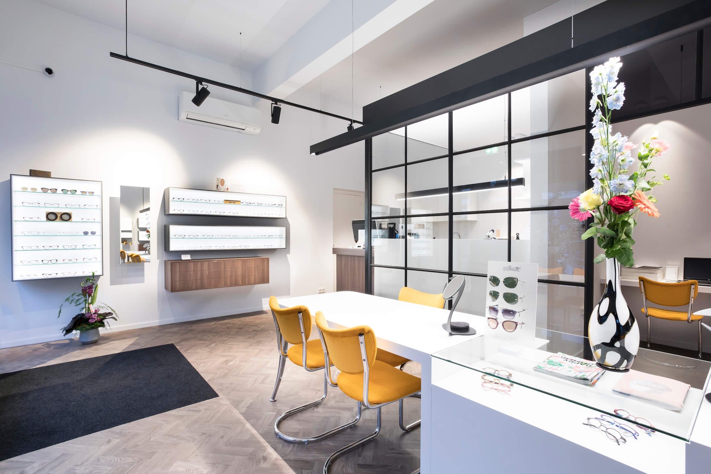Optician interior with lit eyewear displays, yellow chairs and a black steel glass partition